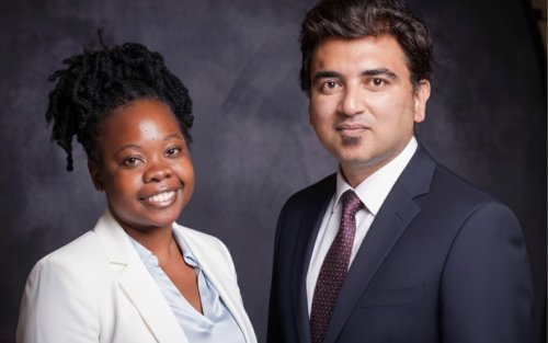 Two professionals, one woman and one man, pose together in formal business attire against a dark studio background.
