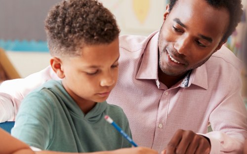 A teacher sits with an elementary age student and points to the student's paper as the student writes.