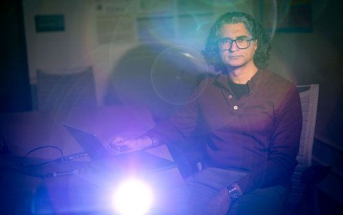A man wearing a maroon shirt and glasses sits in a dark classroom lit by a projector creating ethereal orbs of light across the image.