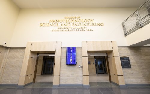 A sign for the College of Nanotechnology, Science, and Engineering hangs above two doorways in a building with light tan and brick walls.