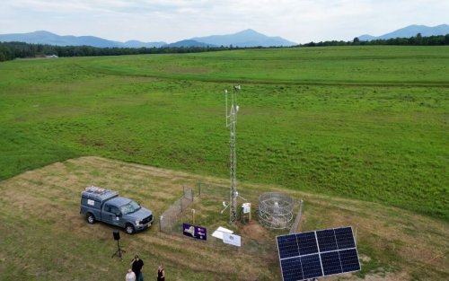 An aerial shot of a fenced in weather station featuring a 10 meter tower and solar panels against a backdrop of a lush green field and mountains in the distance.