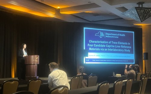 Deanna Luneau stands at a podium, presenting her work. There is a large screen next to her with the title of her presentation written on it: "Characterization of Trace Elements in Four Candidate Caprine Liver Reference Materials via an Interlab study."