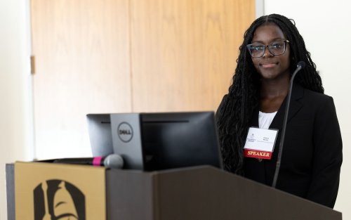 A young African American woman stands at a podium with a gold emblem with the UAlbany logo smiling with a badge that says "speaker"