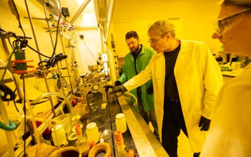 Three people in lab coats stand at a lab bench looking at a piece of equipment in yellow-lit chemistry lab.