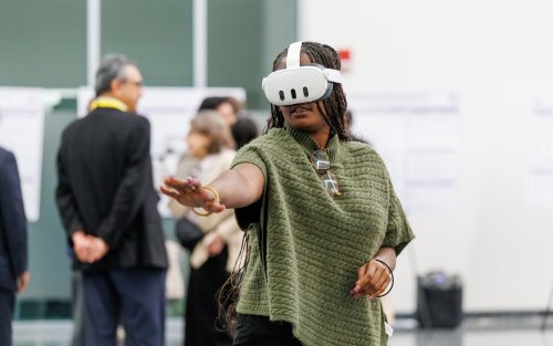 A woman with long braids and a green sweater wears a virtual reality headset and gestures in front of her.