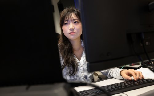 A young woman with dark hair wearing a collared button-down blue and white shirt looks at a pair of monitors in the UAlbany AI for Business Lab