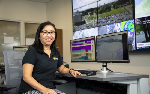 New York State Mesonet Director June Wang smiles in front of computer screens inside the network's operations center.