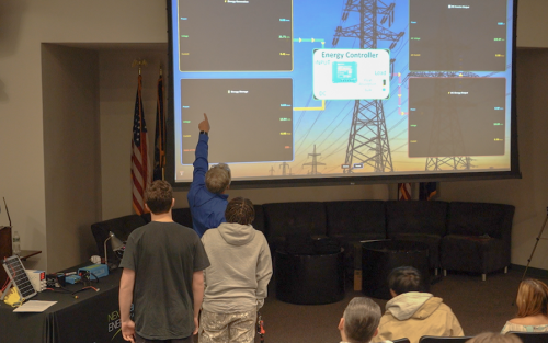 A man at the front of an auditorium points to a screen demonstrating the energy input and output levels of a solar array simulator.