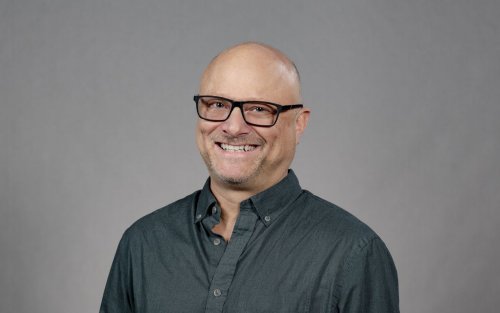 A man wearing a slate button-down shirt and glasses with black rectangular frames smiles for a portrait against a gray backdrop.