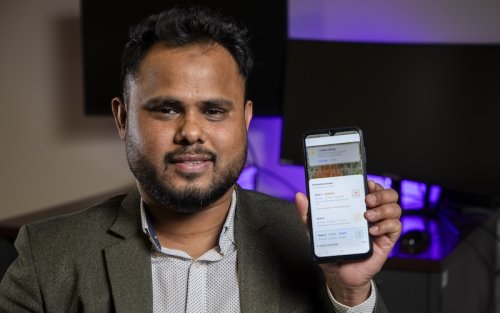 A man in a gray suit jacket holds up a phone with an app on the screen