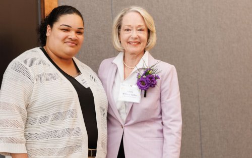 Two smiling women stand together wearing nametags. The older woman has flowers in her lapel.
