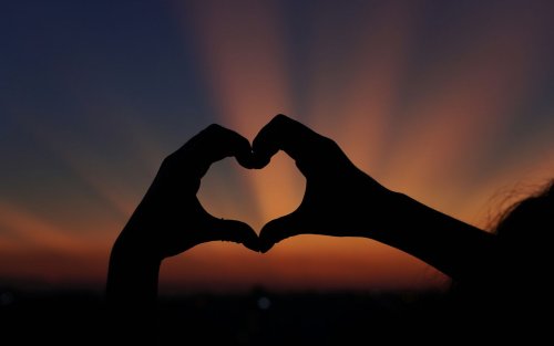 Two hands form a heart shape in front of a sunset, creating a silhouette.