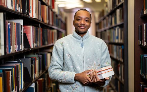 UAlbany student Maurice Burbridge inside the UAlbany library as he stands in front of stacks of books.