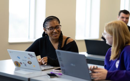 Two students smile and talk while they sit side by side at a desk with their laptops open.