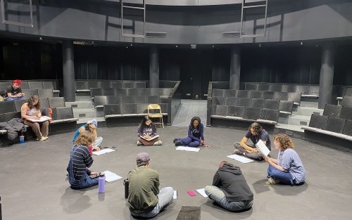 A group of students sit in a theatre-in-the-round with dark seats and walls as they sit in a circle with scripts in front of them.