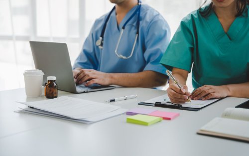 Two people wearing scrubs sit at a white desk. One is working on a laptop, wearing a stethoscope. The other is writing notes on a clipboard. Papers, post-it pads, and several medicine bottles are also on the table.