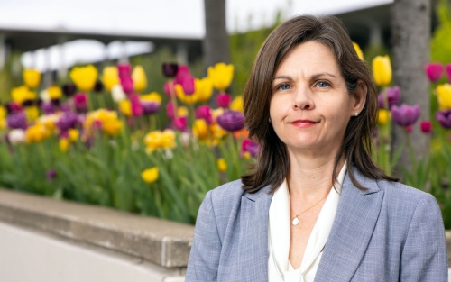 A woman with brown hair wearing a blue-gray blazer and white blouse poses for a portrait in front of a flower bed full of yellow, purple, and magenta tulips in full bloom.