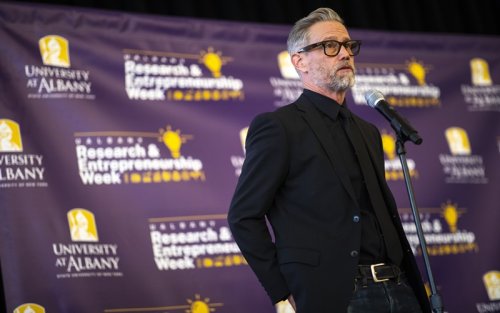 A bearded man with gray/black hair, dark-rimmed glasses and dressed all in black stands at a microphone in front of a purple background that includes the words "University at Albany" and "Research & Entrepreneurship Week"