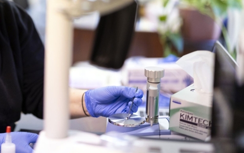 Using a small metal instrument, a person wearing purple latex gloves arranges a tiny amount of a white substance in the center of a round silver platform attached to a piece of lab equipment.