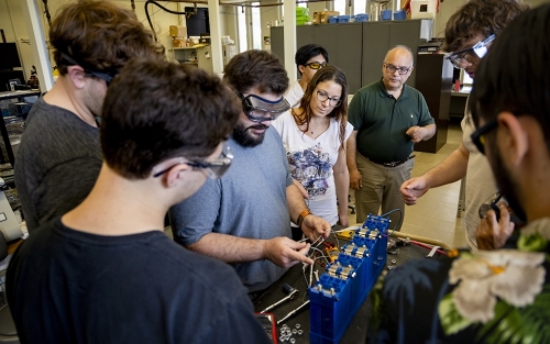 Around half a dozen people in safety glasses gather in a laboratory around a battery as a man holds various wires.