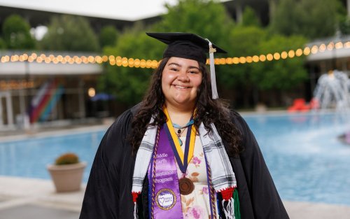 A young woman wearing graduation garb smiles for a portrait in front of Albany’s main fountain. Her cap carries a gray tassel and she is wearing multiple colorful sashes and cords as well as a medal. One of the Sashes us purple satin and says “Middle Earth Peer Assistance Program” with the Middle Earth logo. 