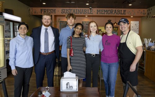 A group of five UAlbany MBA students stand with the owners of fin-your fishmonger inside the store in Guilderland in front of a sign on the wall that says FRESH - SUSTAINABLE - MEMORABLE.