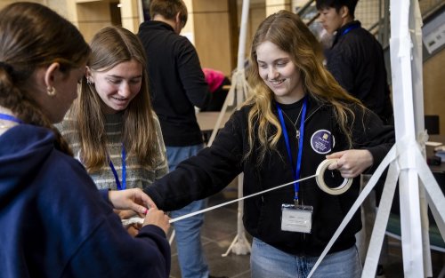 Three smiling teen girls hold onto a spool of masking tape as they attempt to build a freestanding paper tower.