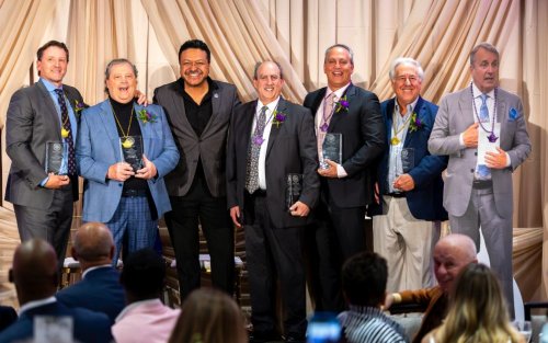 Seven smiling men pose, six of them hold glass awards and wear purple or gold pendants.