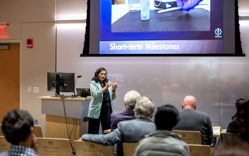 A presenter gestures while speaking to a seated audience at R&E Week 2025. The slide displayed via projector includes the title "Short-term Milestones."