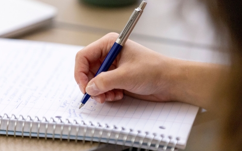 A hand grasps a pen while writing in a spiral bound notebook.