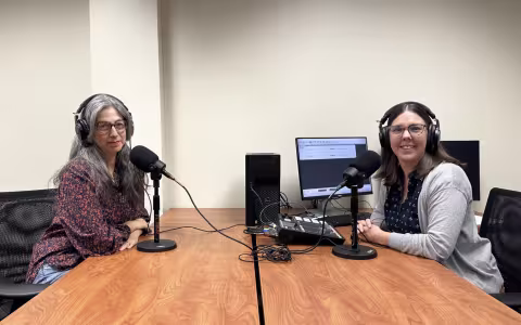 Two women wearing headphones sit in front of microphones at a table next to a computer and recording equipment.