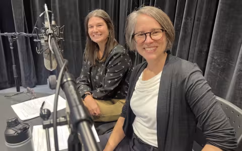 two smiling women sit at a desk with a boom microphone