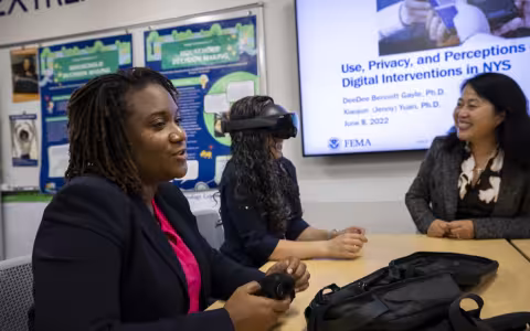 The image features three women seated at a wooden table. A woman wearing a black blazer sits in the foreground beside a woman wearing a black virtual reality headset who is being observed by a smiling woman in a gray blazer sitting across from her. 