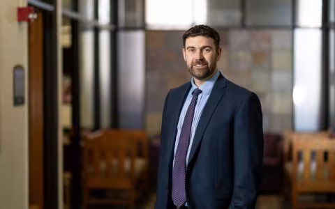 A portrait of a bearded, smiling man standing in a blue suit with a purple tie.