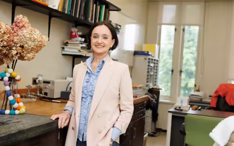 A woman with short brown hair wearing a pale pink blazer and ruffled powder-blue blouse smiles for a portrait in her office.