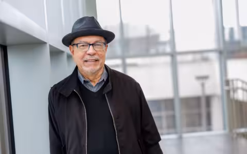 A portrait of a man with eyeglasses in a dark gray hat, black sweater and black jacket leaning against a wall in atrium filled with natural light.