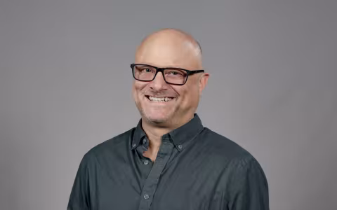 A man wearing a slate button-down shirt and glasses with black rectangular frames smiles for a portrait against a gray backdrop.