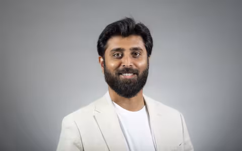 A man with dark brown hair wearing a white T-shirt with white linen blazer smiles for a portrait against a gray backdrop.