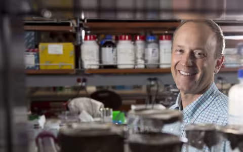 A man smiles for a portrait taken between two shelves crowded with biology lab equipment.