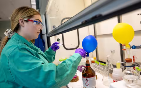 A young woman wearing a green lab coat and purple gloves dips an instrument with a long wire probe into a brown bottle with a small blue balloon attached to its mouth. 