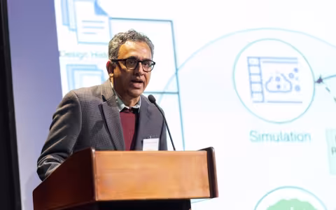 A man with eyeglasses wearing a gray blazer and red sweater stands speaking at a brown lectern with various technical images and the word "Simulation" projected on the screen behind him in blue and green.