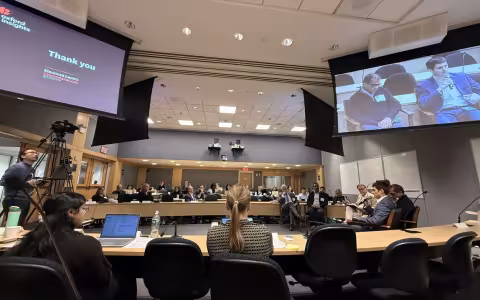 Wide shot of the summit room featuring rows of seated attendees facing a central space in the middle of the room where a group of five men are seated. Four large projection screens hang from the ceiling.