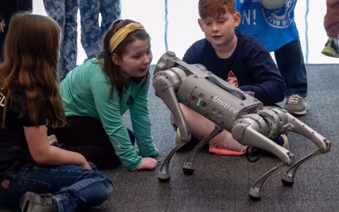 Three children interact with a robotic dog on a gray carpet during UAlbany’s STEM and Nanotechnology Family Day.