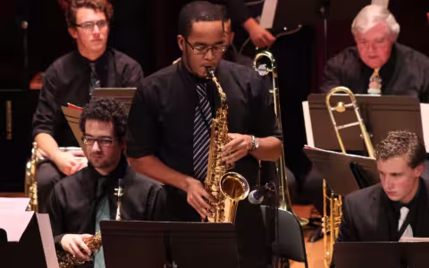A group of musicians wear dark clothing and perform in a jazz ensemble with a saxophone player standing