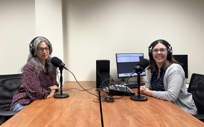 Two women wearing headphones sit in front of microphones at a table next to a computer and recording equipment.