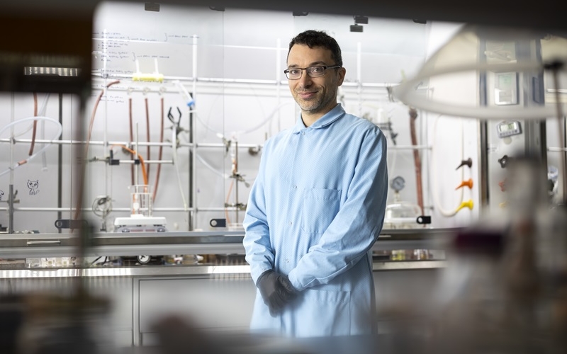 A smiling man in a blue lab coat stands in a chemistry lab surrounded by tubes, workbenches and glassware 
