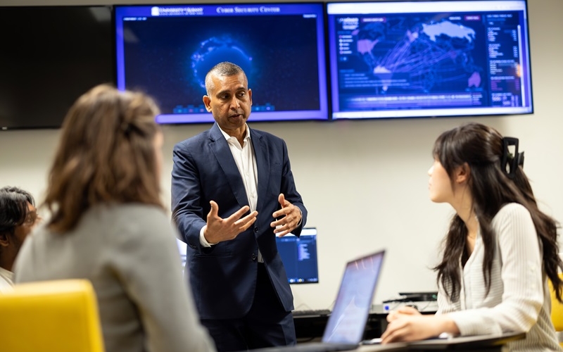 A man in a blue suit stands in front of computer screens gesturing in front of his body while talking to two students.