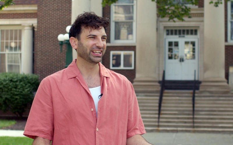 A man wearing a salmon button-down short-sleeved shirt smiles broadly. He is standing in front of a brick building with large cement columns.