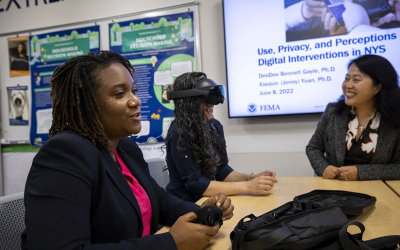 The image features three women seated at a wooden table. A woman wearing a black blazer sits in the foreground beside a woman wearing a black virtual reality headset who is being observed by a smiling woman in a gray blazer sitting across from her. 