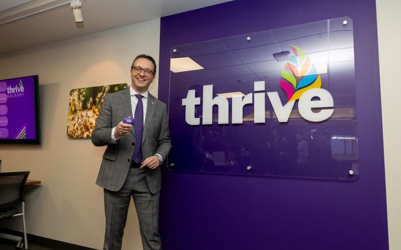 A man in a gray suit smiles while holding up a purple heart in front of a purple wall that says "thrive" in white letters.